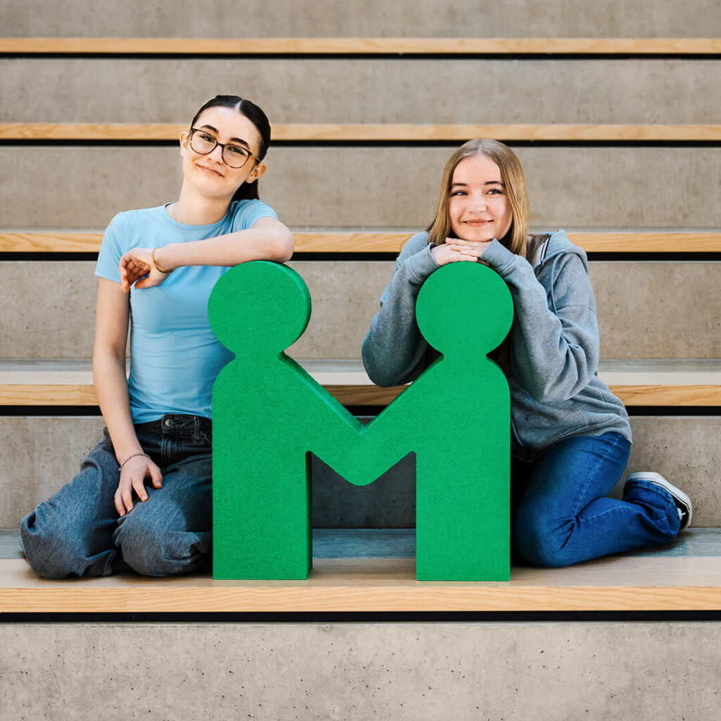 Schuelerinnen einer Mentor Kooperationsschule sitzen auf einer Treppe mit der Logo Figur